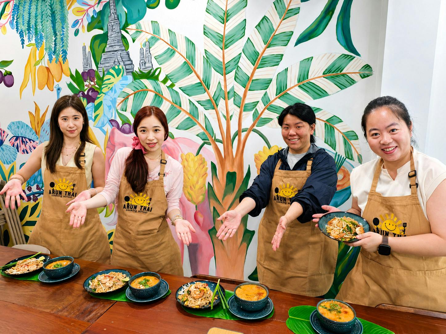 Three people wearing aprons stand behind a table with dishes of food, smiling and gesturing towards the dishes.