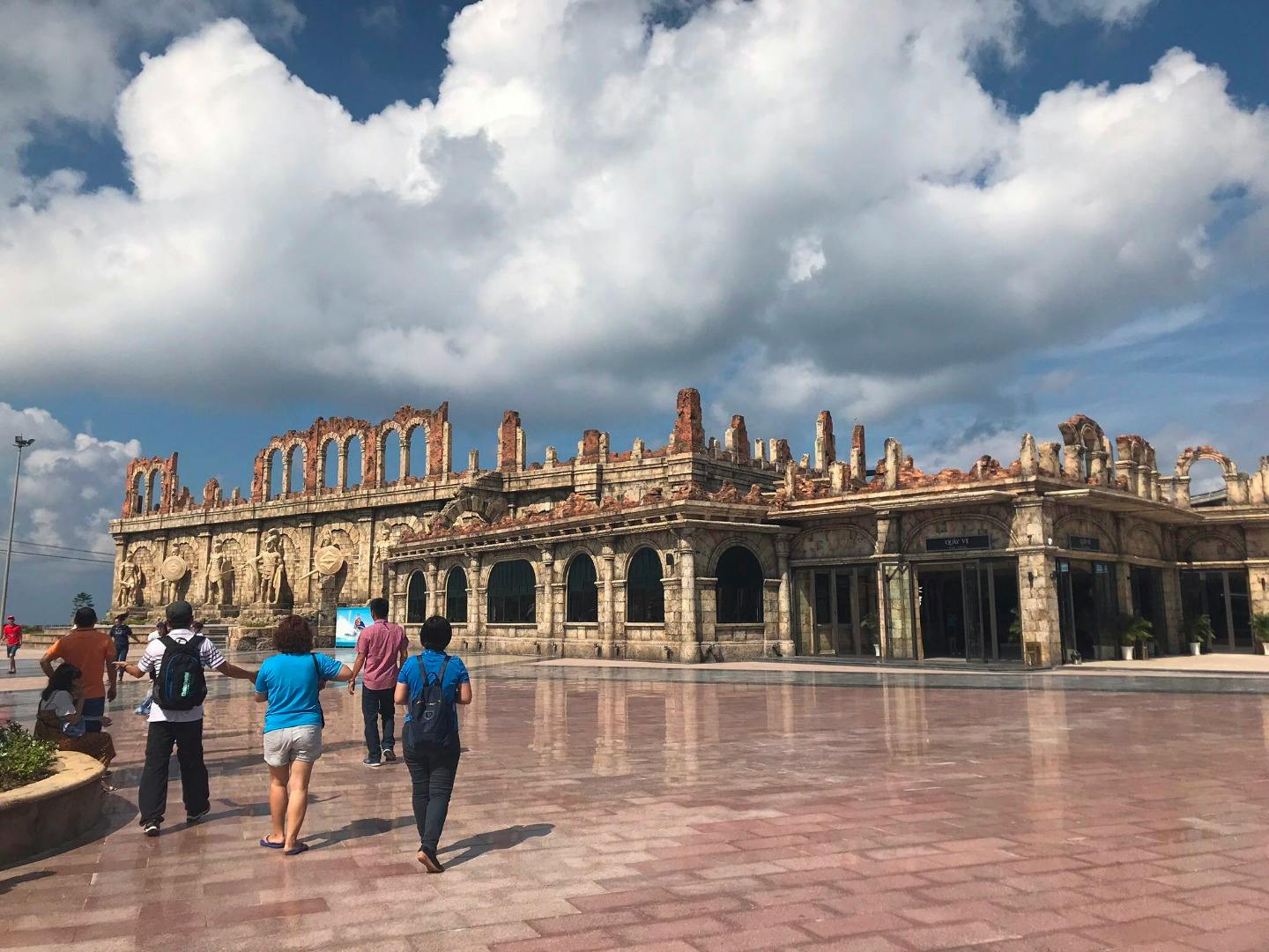 People walking on a plaza near a large, ancient-style stone building with arches under a partly cloudy sky.
