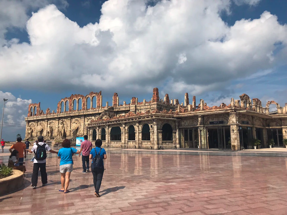 A group of people walking towards a large stone building with arches and ruins under a partly cloudy sky.