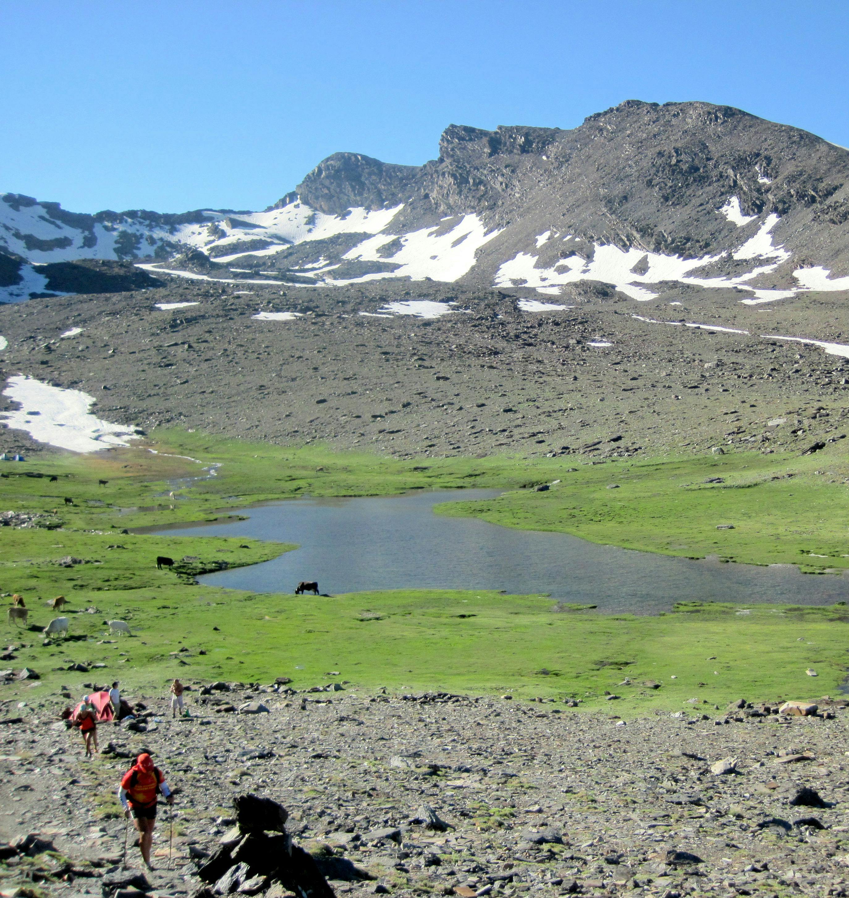 Hikers in the foreground walk toward a small lake surrounded by rocky terrain and patches of snow under a clear blue sky.