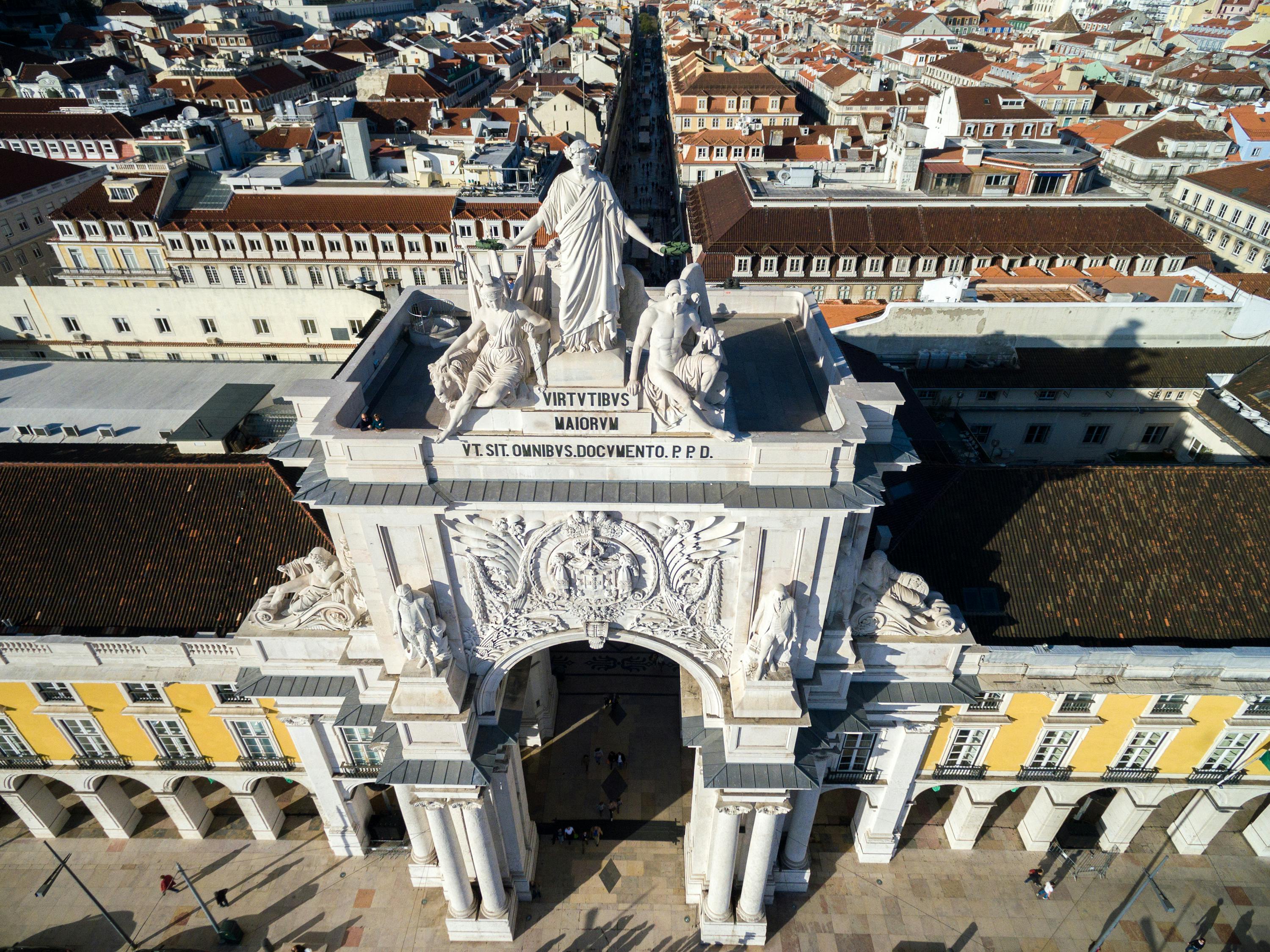 Flygfoto över Rua Augusta Arch i Lissabon, Portugal, med statyer på toppen och en livlig stadsbild med terrakottatak i bakgrunden.