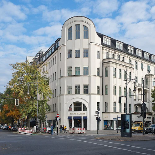 Gatuhörn med en stor vit byggnad med "Ralph Lauren" -skyltar, träd, fotgängare, bilar och en delvis molnig himmel.