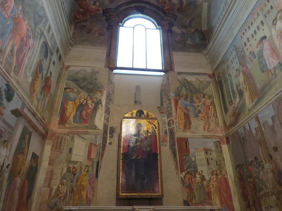 Interior of a chapel featuring a stained glass window above painted murals on walls and a central icon of religious figures.