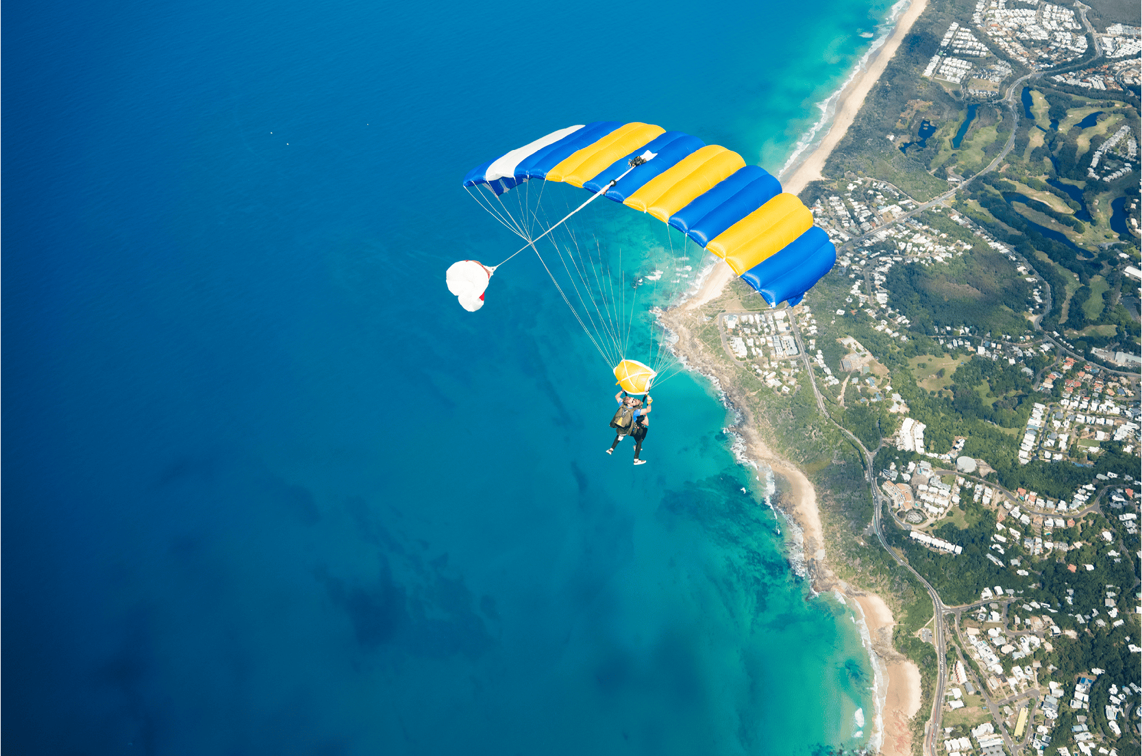 Skydiver with a yellow and blue parachute descending over a coastal area with blue ocean and beaches below.