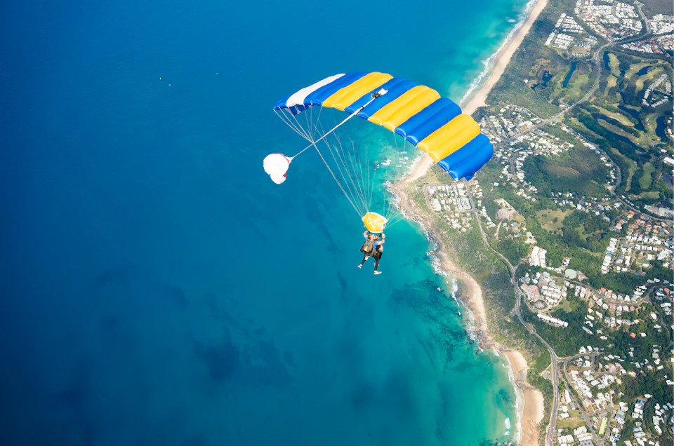 Skydiver with a yellow and blue parachute descending over a coastal area with blue ocean and beaches below.