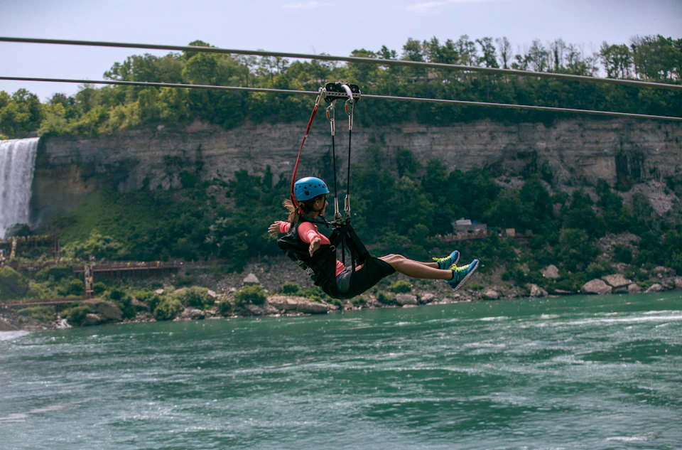 A person wearing a helmet and harness zip-lining over a river with a forested cliff in the background.
