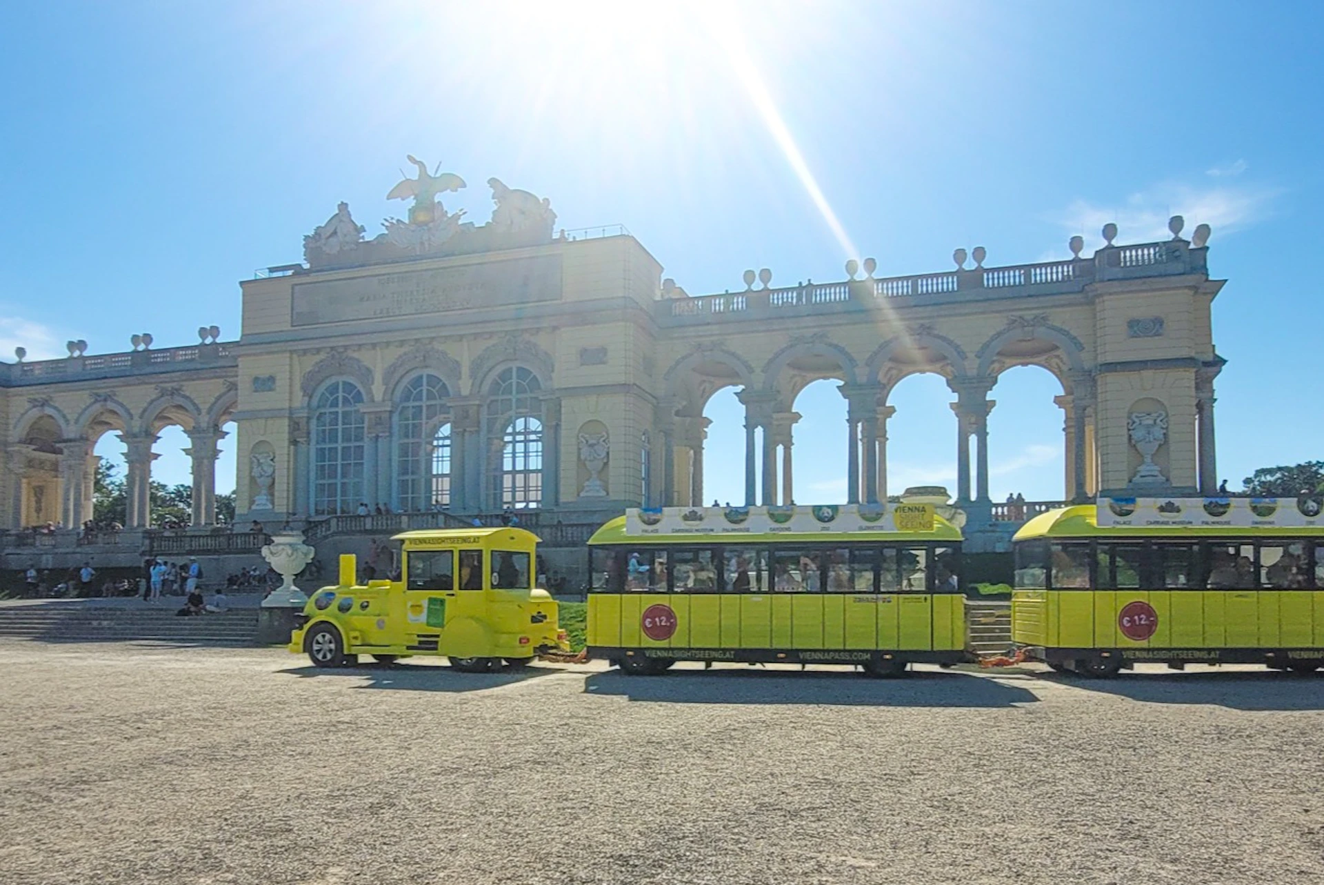 A yellow tourist train in front of the Gloriette structure at Schönbrunn Palace under a sunny sky.