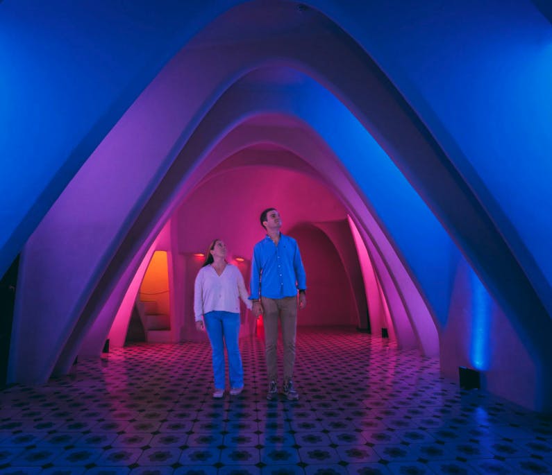A couple walks under arched structures in a room illuminated by blue and pink lights with patterned flooring.