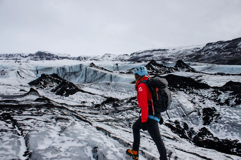 A person wearing a red jacket and helmet hiking on a snowy glacier terrain under a cloudy sky.