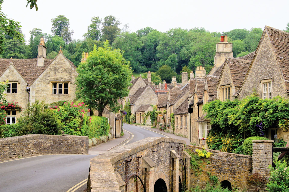 A scenic village with stone houses, a bridge in the foreground, and lush greenery in the background.