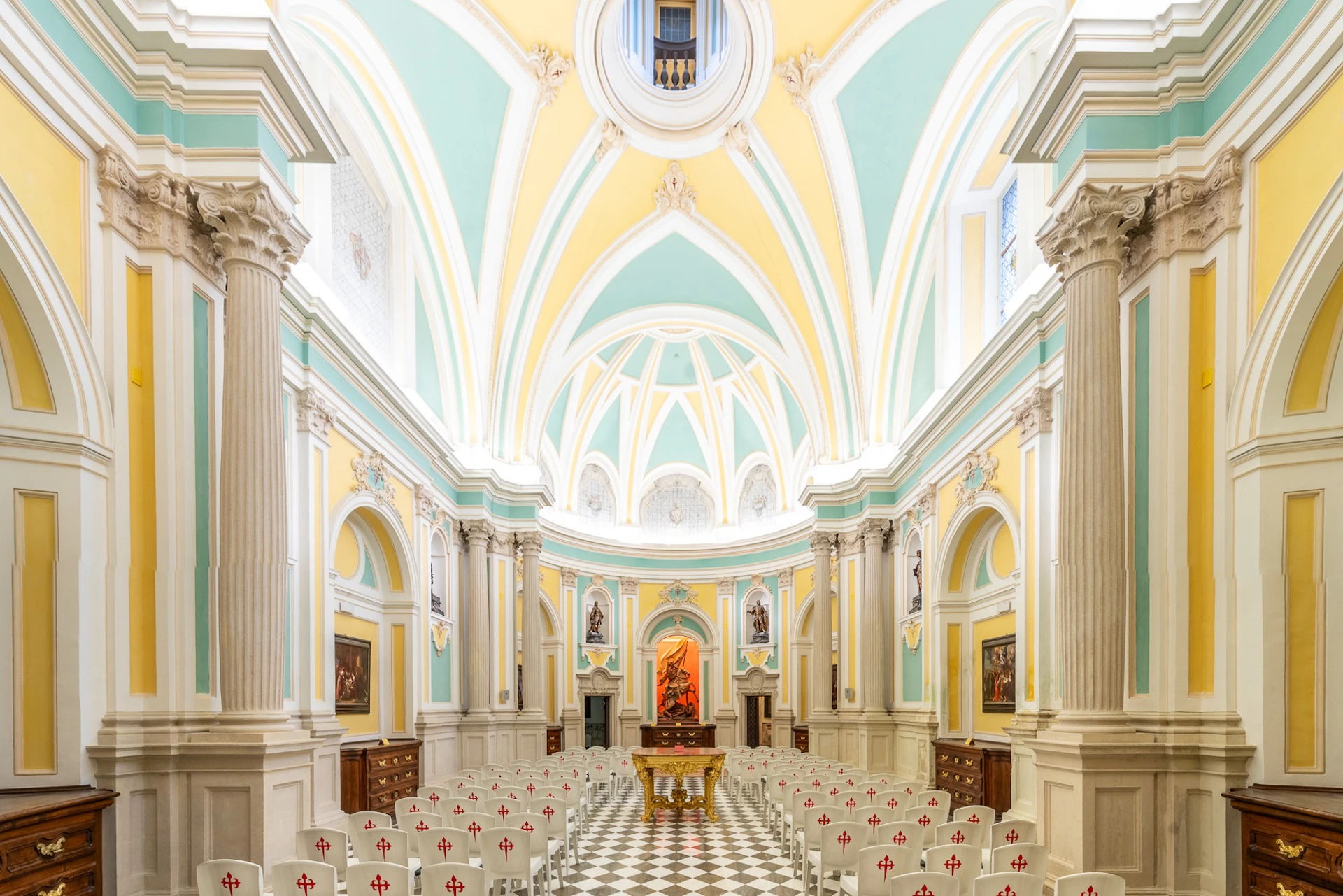 A brightly lit church interior with arched ceilings, decorative columns, and rows of chairs facing an altar.