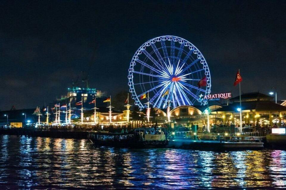 Nighttime view of a lit Ferris wheel and buildings reflecting on a river, with "Asiatique" signage visible.