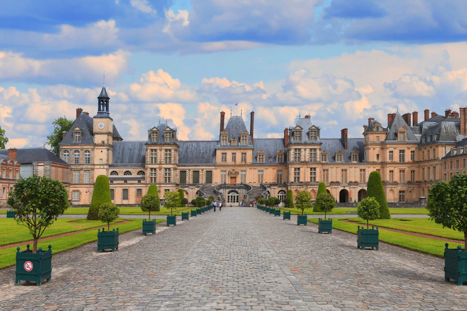 A symmetrical palace with a cobblestone path leading to its entrance, surrounded by manicured lawns and trees under a blue sky.