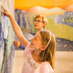 Två barn ler och rör vid en färgglad mosaikvägg, med fokus på dess struktur och mönster.