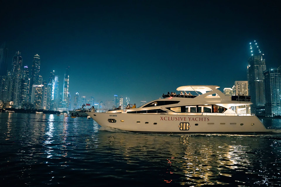 Luxury yacht "XCLUSIVE YACHTS" on water at night, city skyline with illuminated buildings in background.