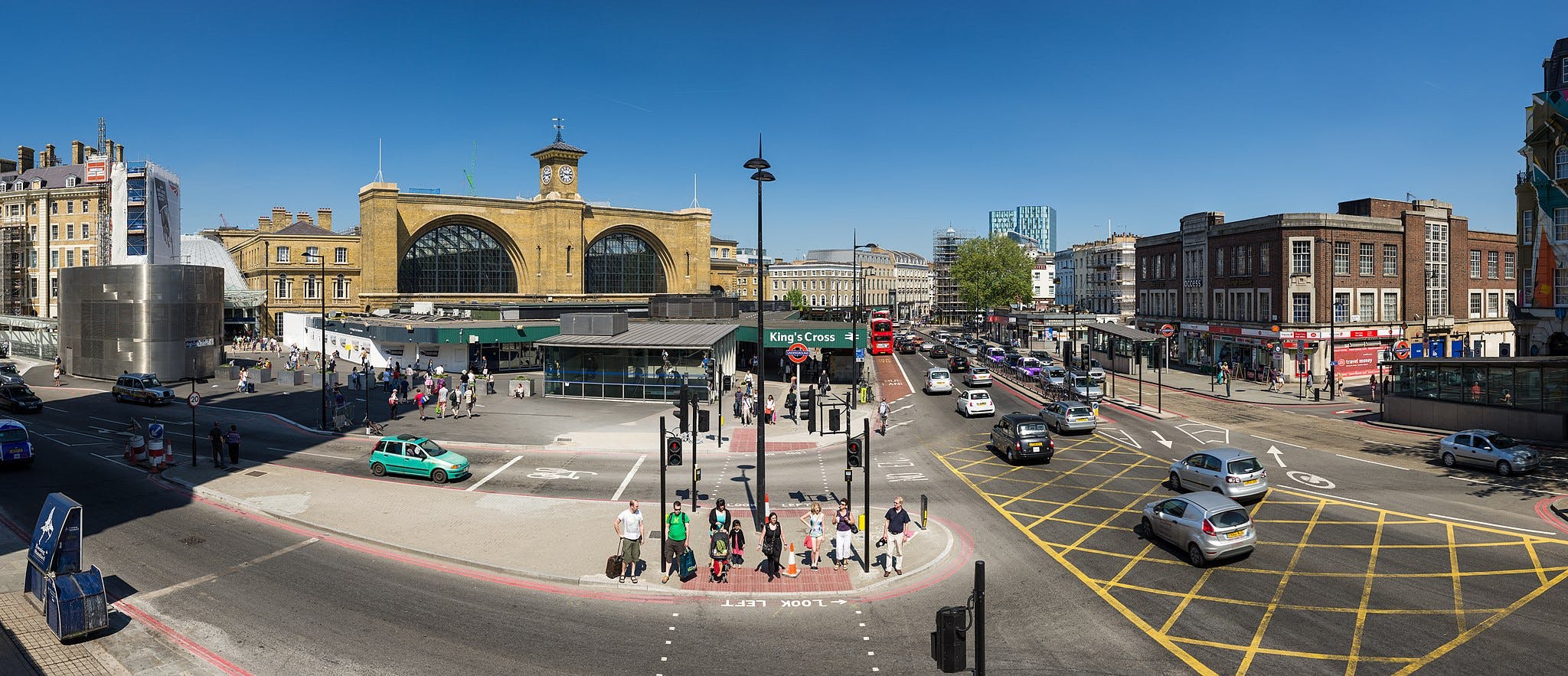 Busy urban intersection with pedestrians, cars, and a red double-decker bus near a large brick building and King's Cross Station.