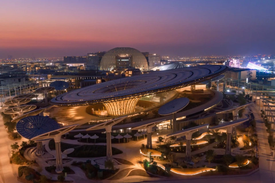 A futuristic architectural structure with a large circular canopy, surrounded by illuminated pathways at dusk.