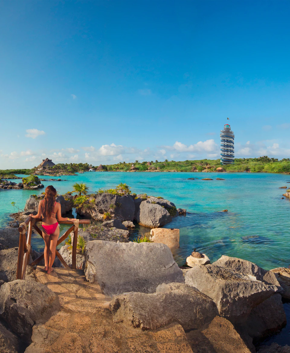 A woman in a pink bikini stands on a rocky path overlooking a clear turquoise lagoon with a unique spiral tower in the background.