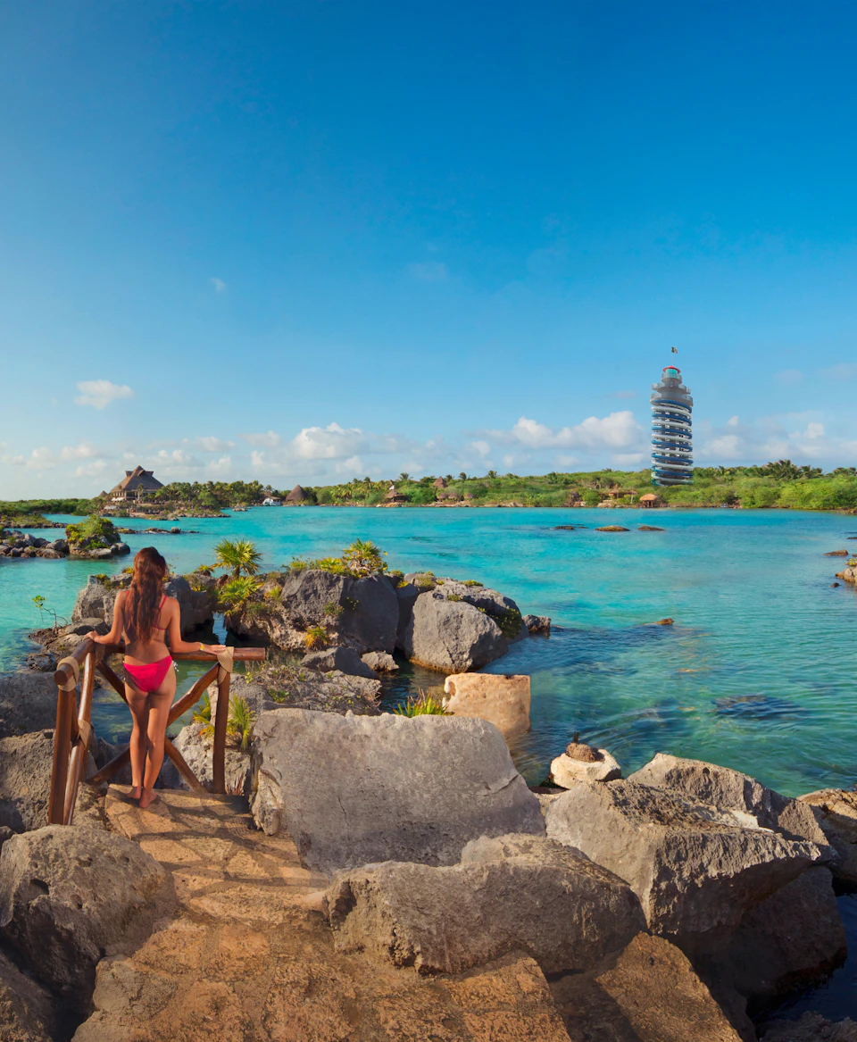 A woman in a pink bikini stands on a rocky path overlooking a clear turquoise lagoon with a unique spiral tower in the background.