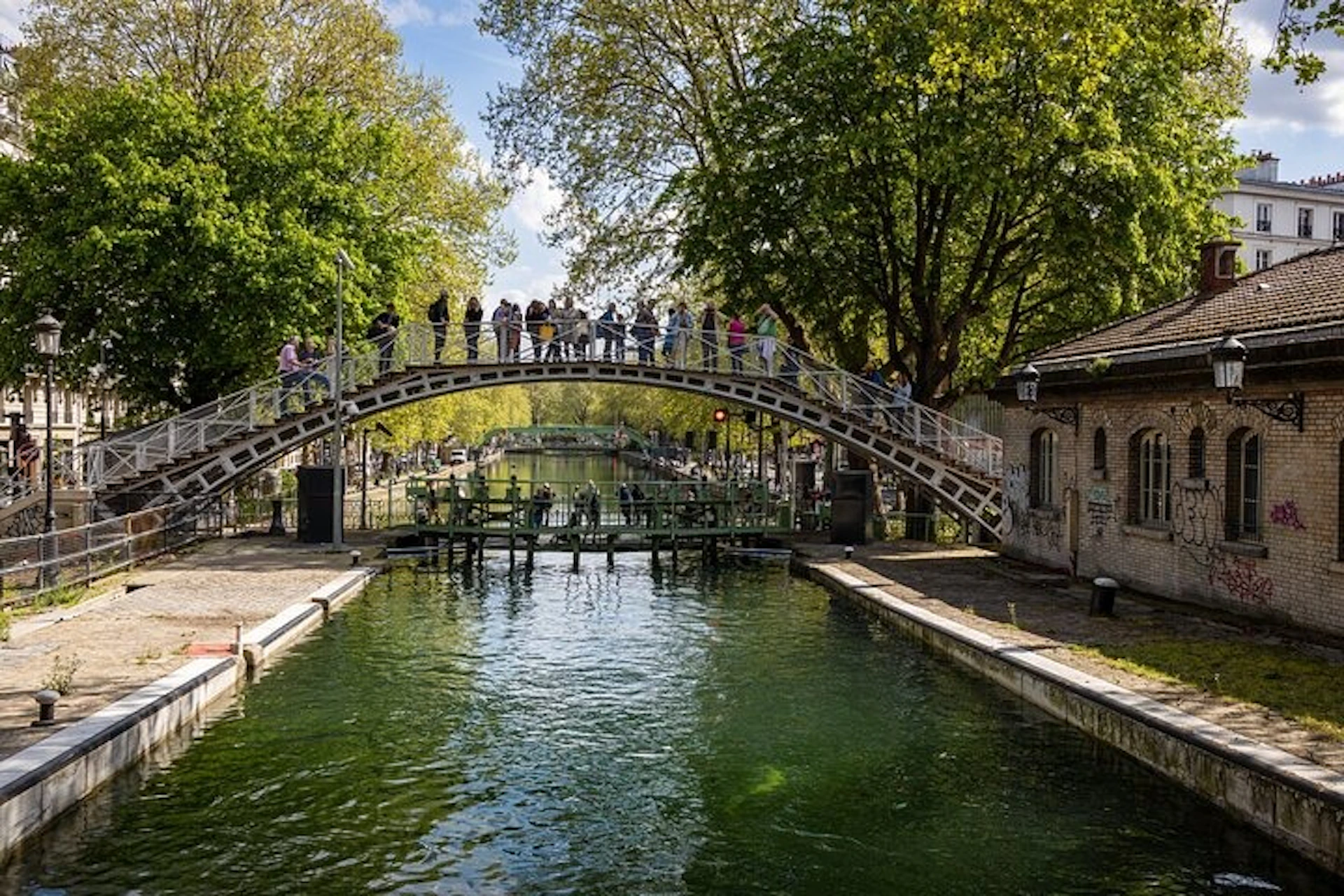 A pedestrian bridge crosses a canal surrounded by green trees. People are walking and standing on the bridge.