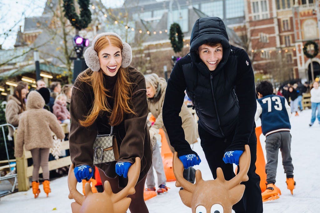 Δύο άνθρωποι κάνουν πατινάζ στον πάγο με ενθουσιασμό, κρατώντας στηρίγματα σε σχήμα ταράνδου, περιτριγυρισμένοι από άλλους που κάνουν πατινάζ σε ένα εορταστικό χειμωνιάτικο σκηνικό.