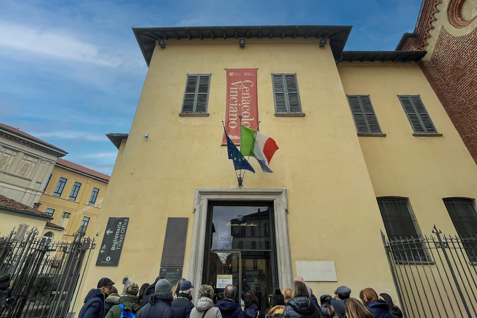 A group of people gathered outside a beige building with a sign reading 'Cenacolo Vinciano' and flags above the entrance.