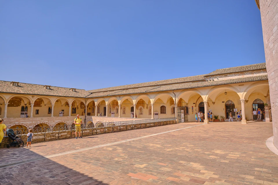 A courtyard with a rectangular building featuring arches and a tiled roof, with people walking and standing on the paved ground.