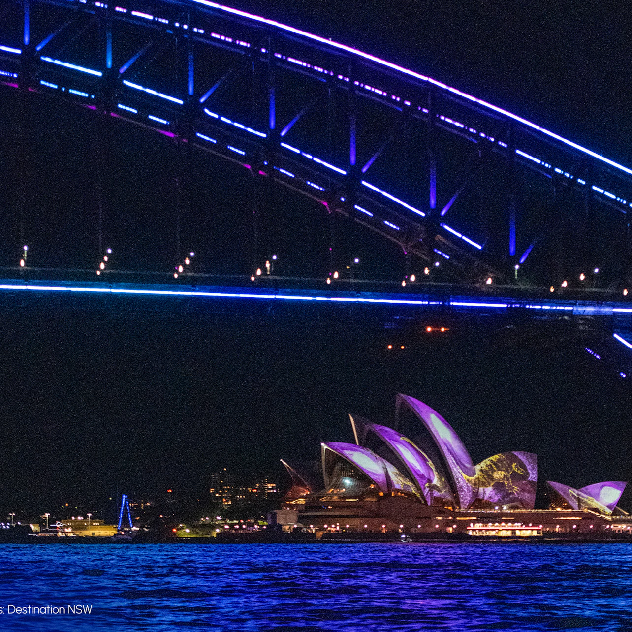 The Sydney Harbour Bridge lit with colorful lights at night, with the Sydney Opera House illuminated in the background.