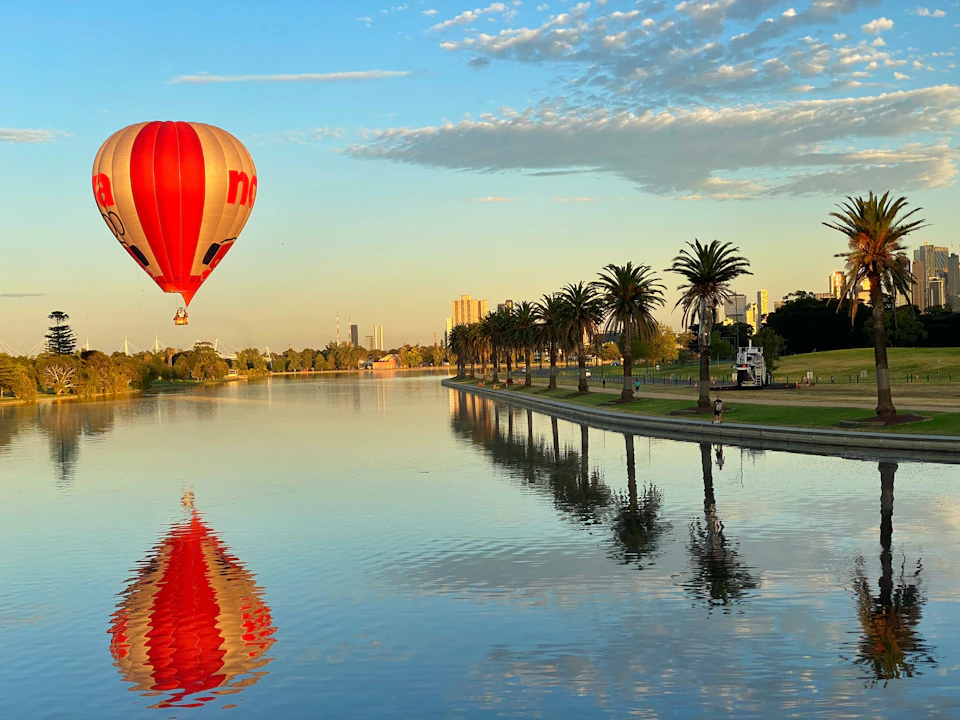 A hot air balloon floats over a calm lake lined with palm trees. The sky is clear with some clouds, and the reflection is visible in the water.