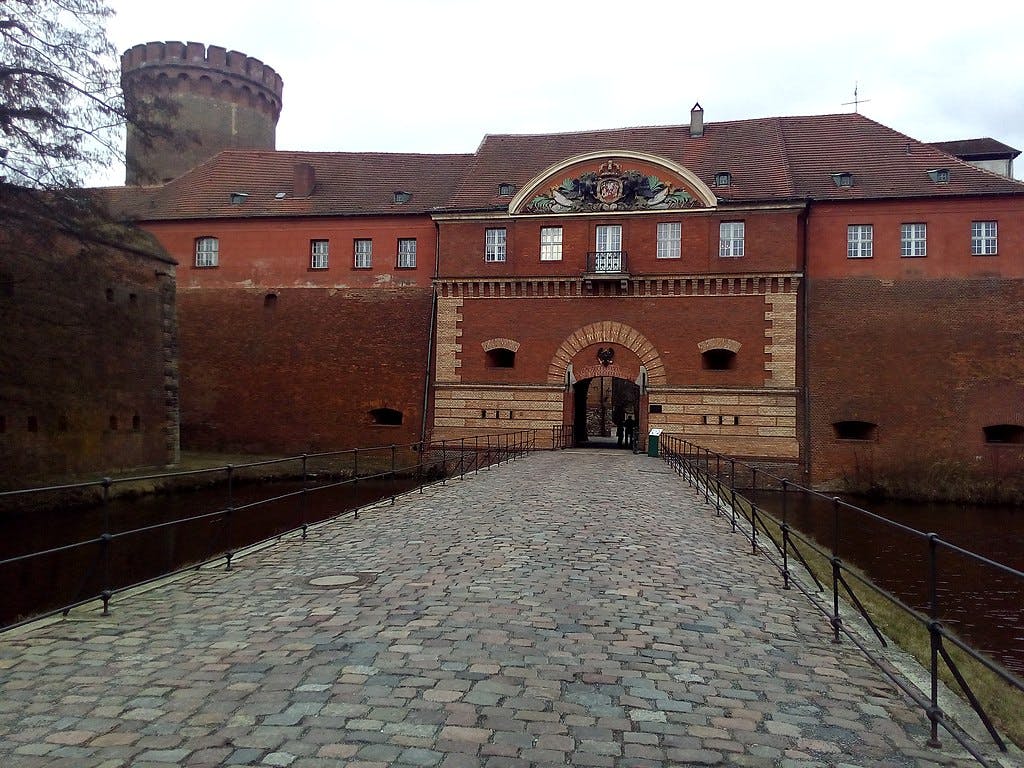 Cobblestone bridge leading to an arched entrance of a red-brick building with a tower, moat, and decorative crest above the arch.