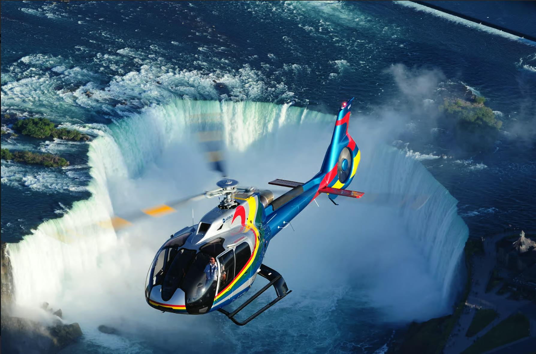 A colorful helicopter flies near the misty edge of a large waterfall with powerful water flow in the background.