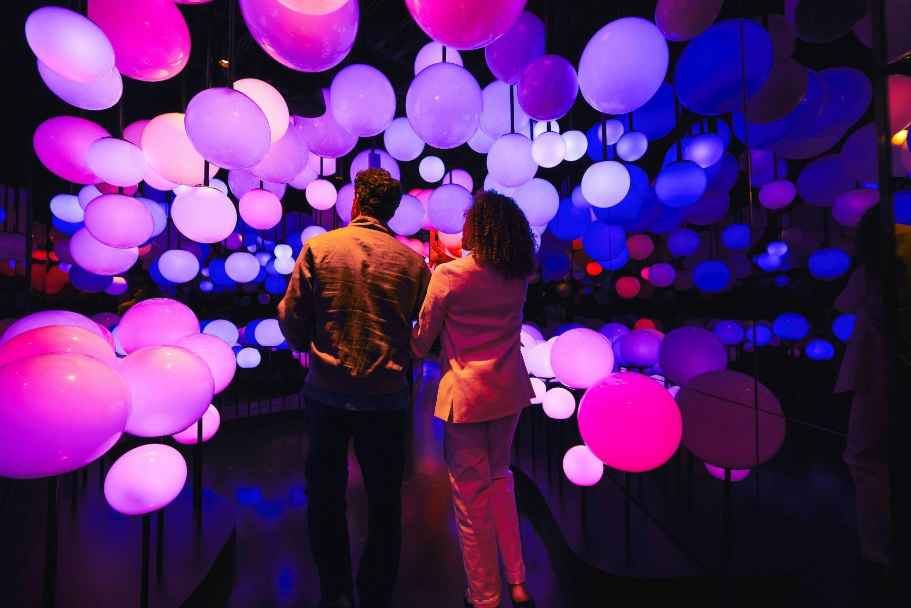 Two people walk through a colorful installation of glowing spheres in a dark room with vivid purple and pink lighting.