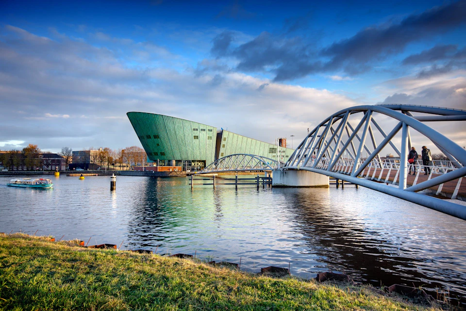 Modern green building next to a river, with a curved white bridge spanning the water and a grassy bank in the foreground.