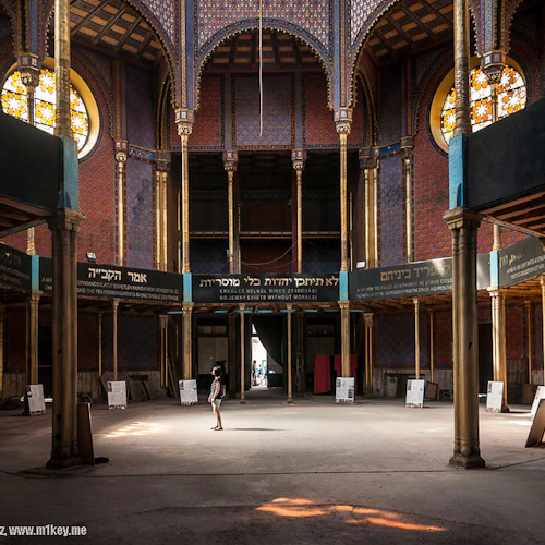 En person står i en stor, utsmyckad hall med glasmålningar, välvda tak och hebreisk text på banderoller.
