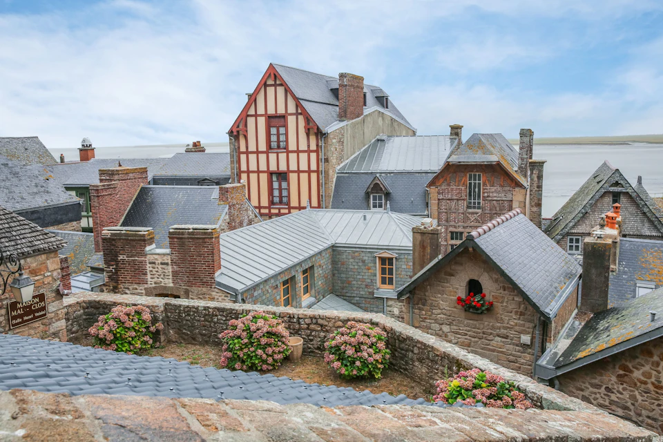 Rooftops of a cluster of historic buildings with stone and brick construction, flowers in the foreground, under a partly cloudy sky.