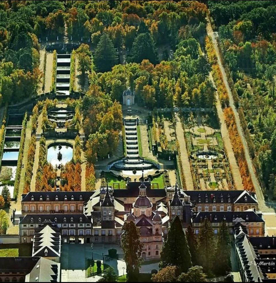 Aerial view of a large palace with symmetrical gardens, fountains, and tree-lined pathways in an autumn landscape.