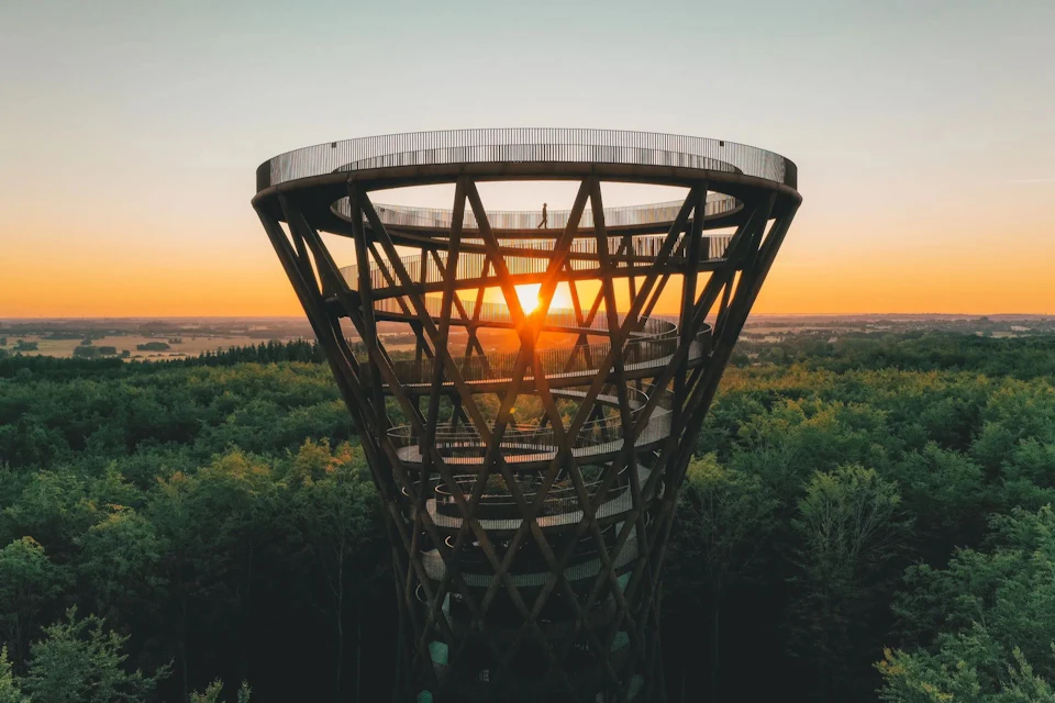 A spiral observation tower with a lattice structure, set against a sunset over a forested landscape.