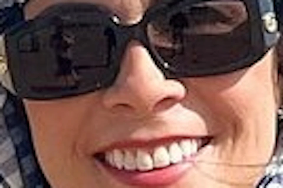 A woman wearing sunglasses and a headscarf smiles while standing next to a camel, with pyramids visible in the background.