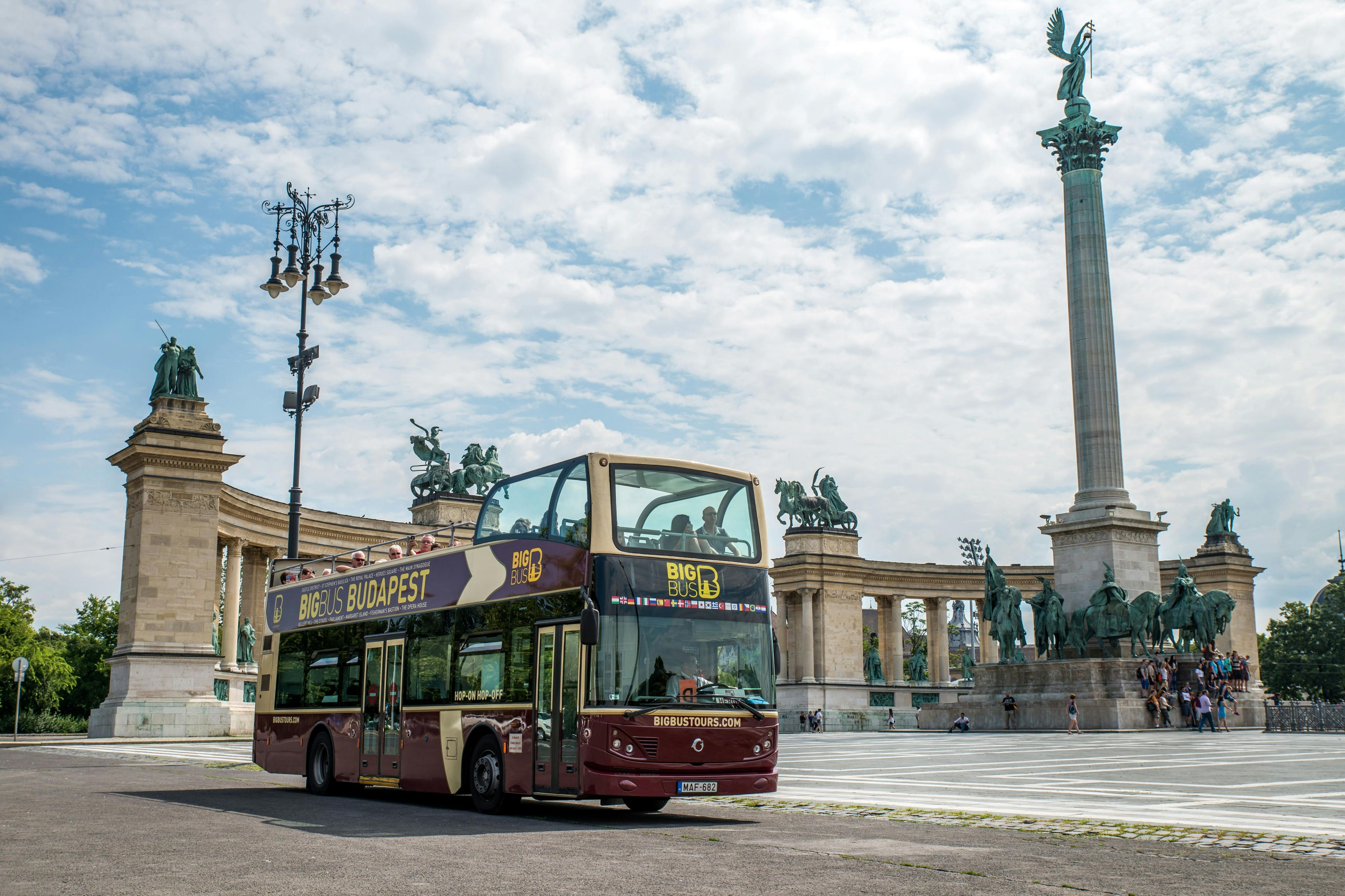 Un autobús turístic de dos pisos a Budapest, prop de la Plaça dels Herois, amb estàtues i un monument alt al fons.