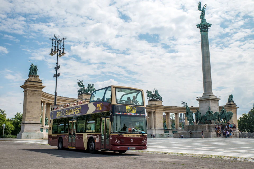 A double-decker sightseeing bus in Budapest near Heroes' Square, with statues and a tall monument in the background.