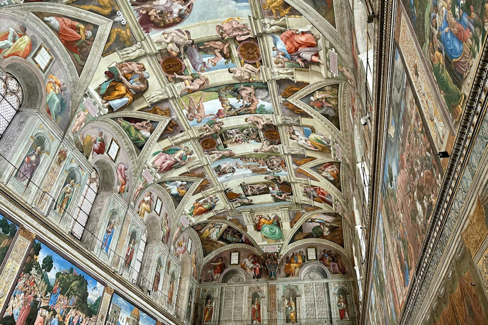 A view of the Sistine Chapel ceiling and walls adorned with detailed frescoes depicting scenes from the Bible.