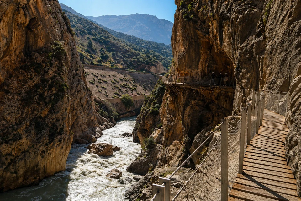 A narrow wooden walkway along the cliff face above a river in a canyon, with views of hills and a blue sky in the background.