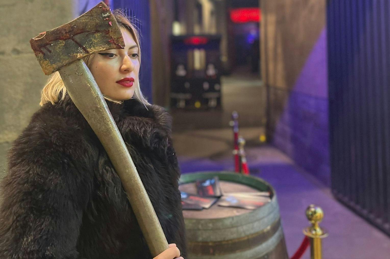 身穿黑色毛皮大衣、头发长长的金发女郎，手里拿着一把风化的大斧头。 背景有一个木桶和城市灯光。