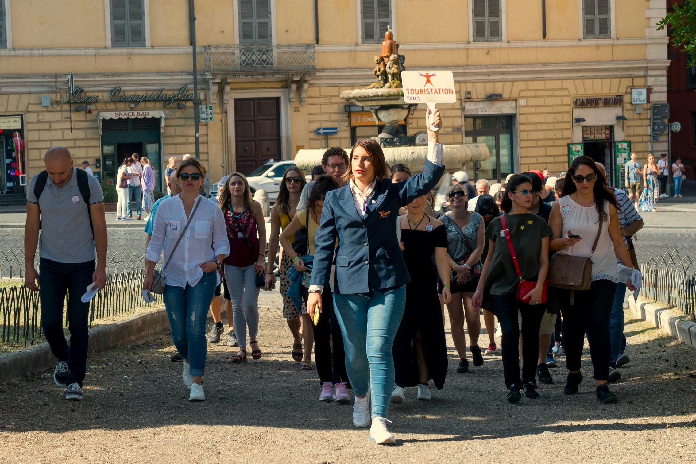 A tour guide holding a sign leads a group of people through a sunny urban area with a fountain and buildings in the background.