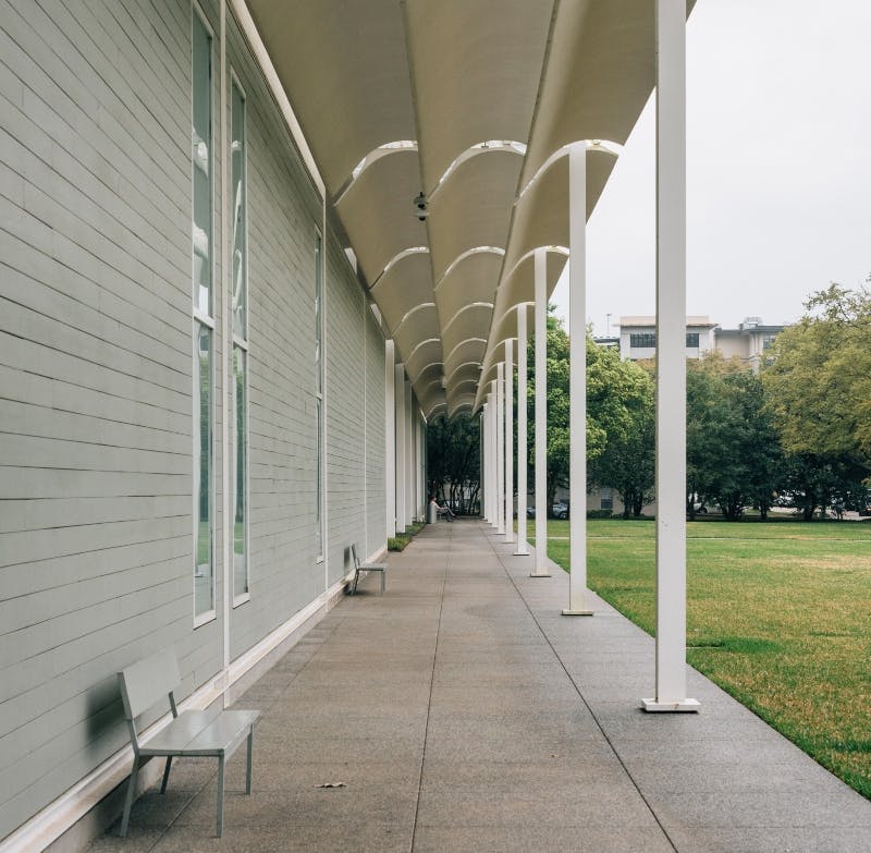 커다란 창문이 있는 건물에 인접한 현대적인 돌출부가 있는 긴 산책로. 왼쪽에는 빈 의자가 놓여 있습니다.