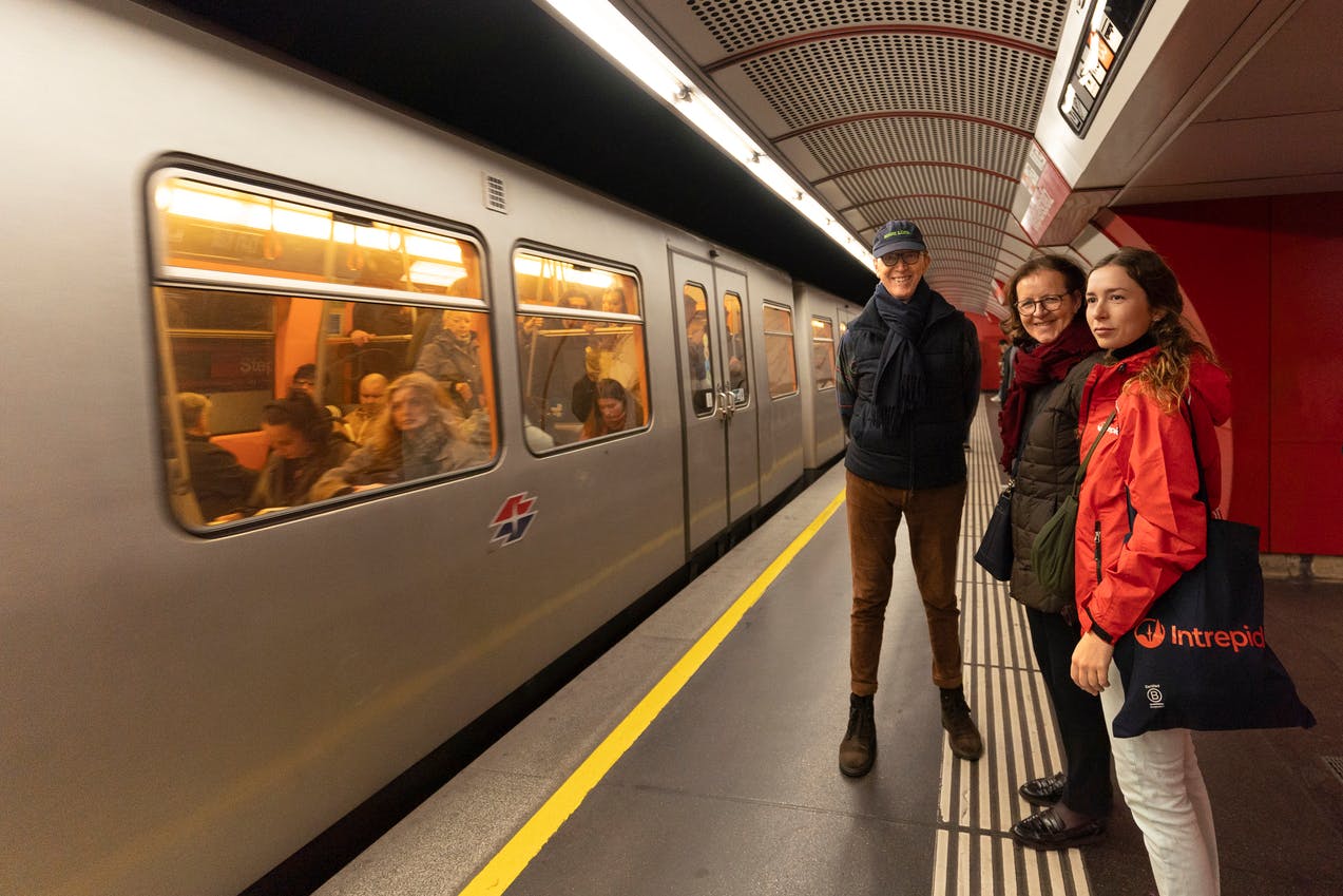 Três pessoas numa plataforma de metro ao lado de um comboio parado, com passageiros visíveis no interior e paredes vermelhas ao fundo.