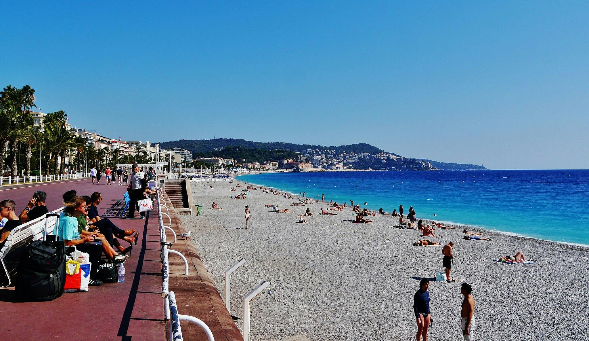 People relaxing on benches along a coastal promenade; others sunbathing and swimming on the adjacent rocky beach under a clear blue sky.