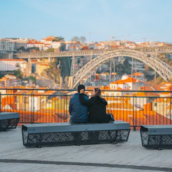 Two people sitting on a bench overlooking a cityscape with a bridge and red-roofed buildings under a blue sky.