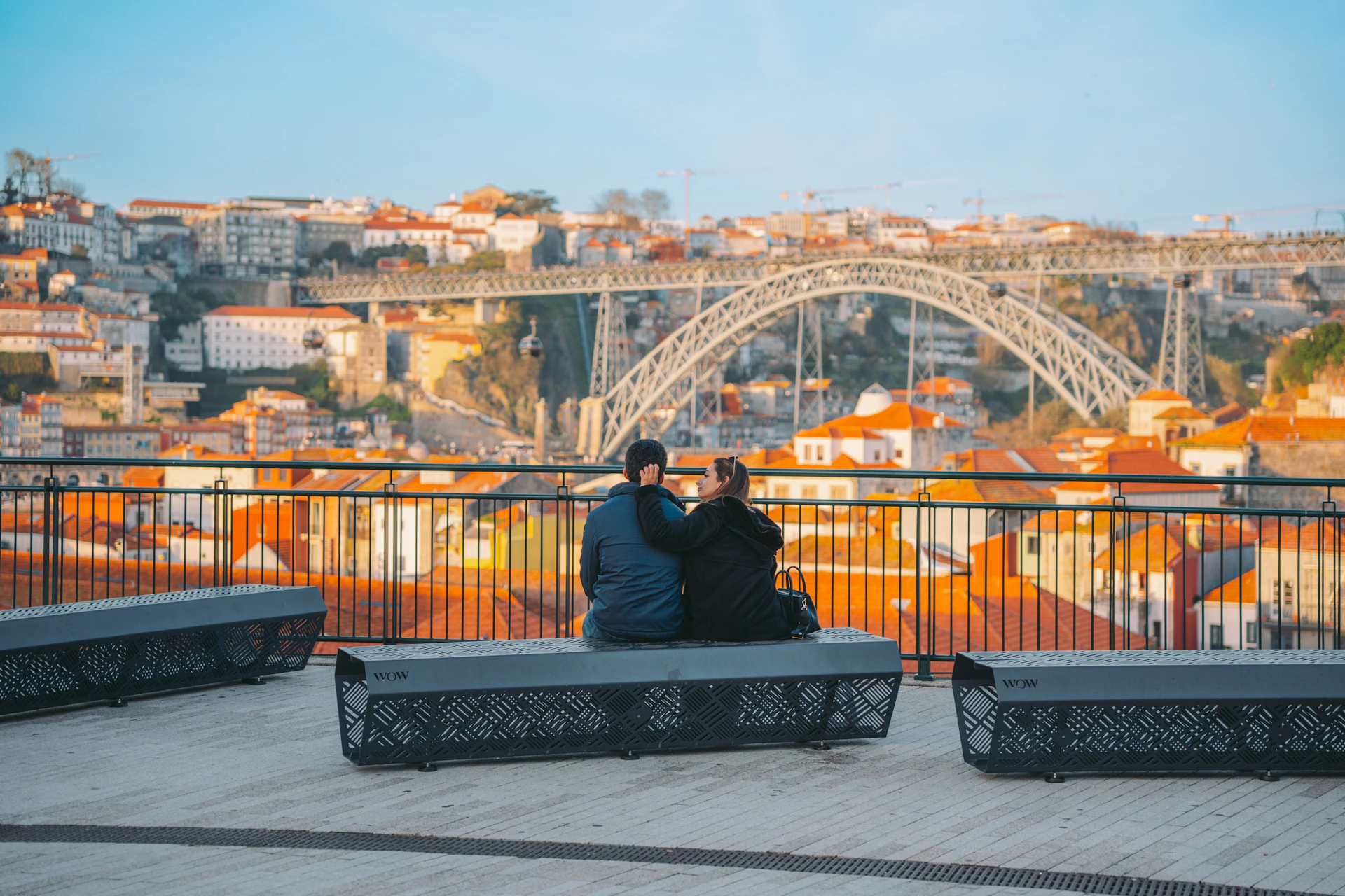 Two people sitting on a bench overlooking a cityscape with a bridge and red-roofed buildings under a blue sky.