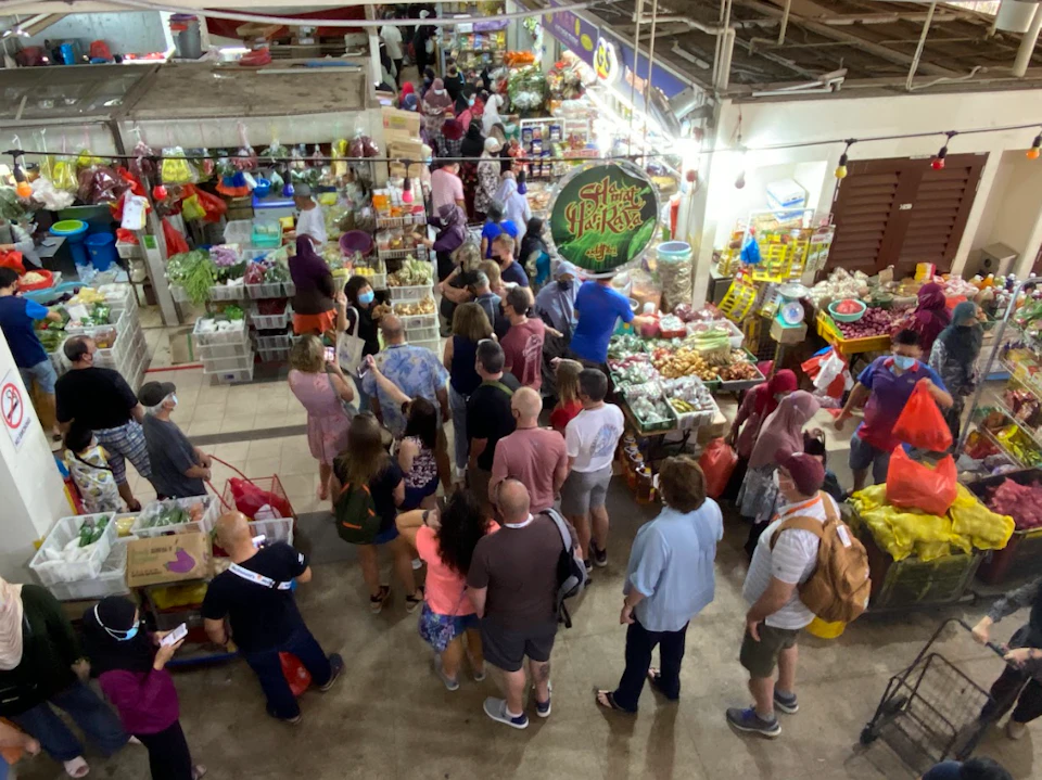 Crowded indoor market with people standing in a line, surrounded by various stalls selling fresh produce and goods.